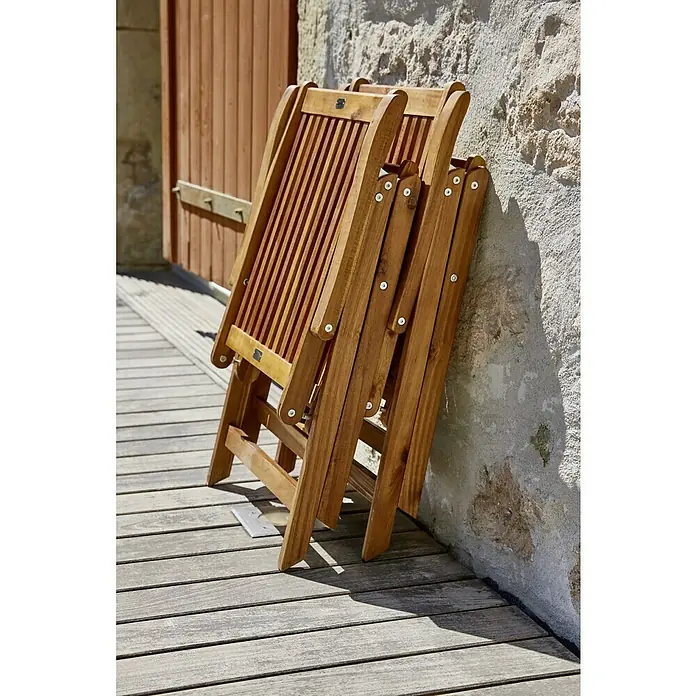 Sunfun Gartenmöbel-Set Diana/Trentino - 6 -tlg., Akazie/Aluminium/Polyester, Braun/Chili - Zwei zusammengeklappte Holzgartenstühle lehnen an einer Wand und warten auf ihren Einsatz. Die Stühle sind aus hellem Holz gefertigt.