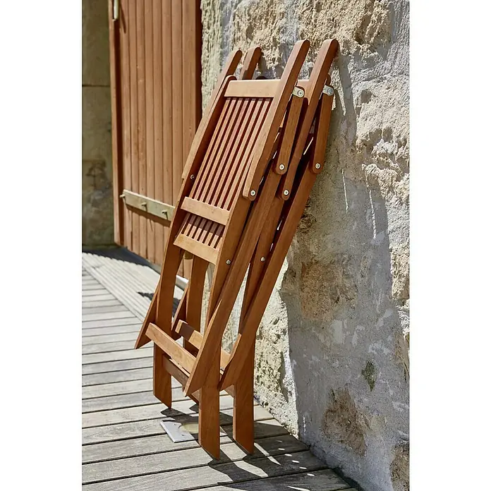 Sunfun Gartenmöbel-Set Diana - 5 -tlg., Akazienholz, Niederlehner mit Armlehne, Tischplatte ausziehbar - Aufnahme eines zusammengeklappten Holzstuhls an einer rustikalen Wand, der im Freien steht und im Sonnenlicht ein warmes, einladendes Ambiente versprüht.