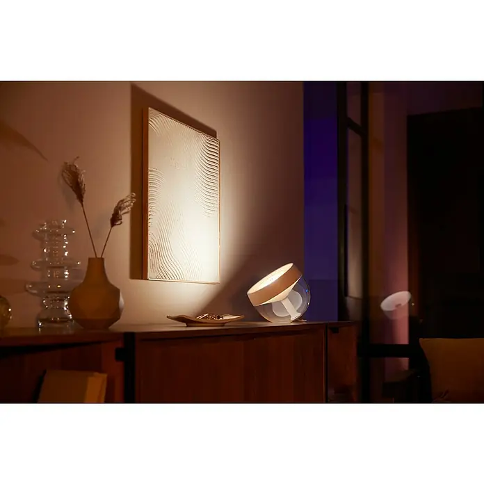Philips Hue LED-Tischleuchte Iris - Weiß/Transparent, Höhe: 19,4 cm - Ein stimmungsvolles Ambiente mit der Philips Hue Lampe, die warmes Licht auf ein Kunstwerk wirft.