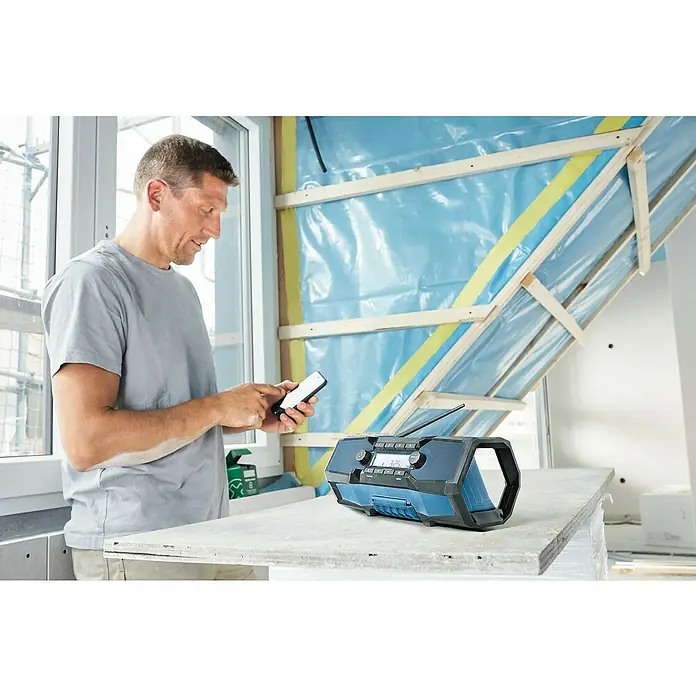 Bosch Professional AMPShare 18V Radio de batería para la obra GPB 18V-2 C - 18 V, Sin batería, 87,5 - 108 MHz (VHF/FM) - Un hombre con un teléfono inteligente está de pie junto a una radio de obra Bosch, robusta y resistente a la intemperie, ideal para uso en exteriores.