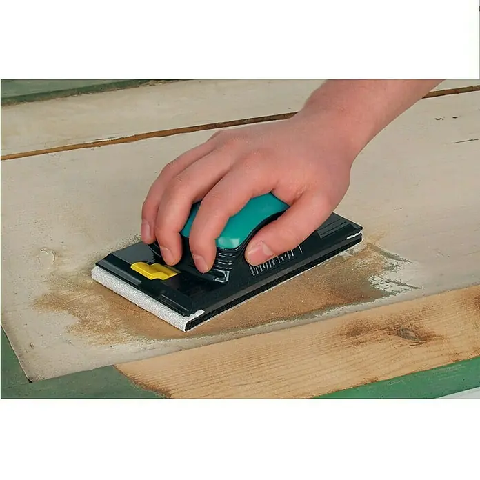 Wolfcraft Lijadora manual - Plástico - Mano con una lijadora manual sobre una tabla de madera con pintura verde. La herramienta tiene un mango verde y una base negra.