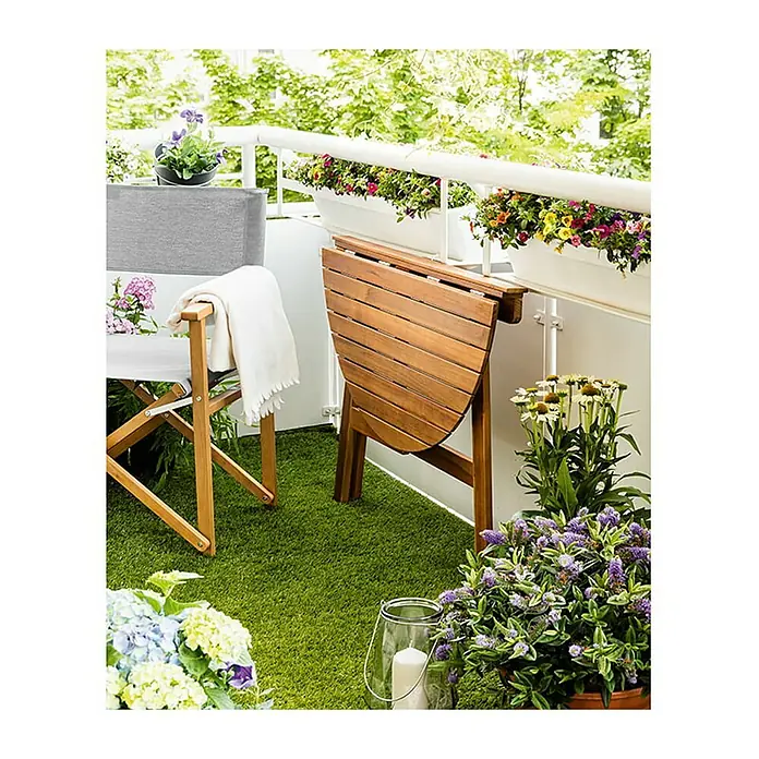 Sunfun Diana Mesa de balcón - L x An x Al: 100 x 50 x 75 cm, Acacia, Abatible, Curvado, Marrón natural - Mesa plegable de madera en un balcón. La mesa está medio plegada y está al lado de una silla con una manta. Jardineras adornan la barandilla.