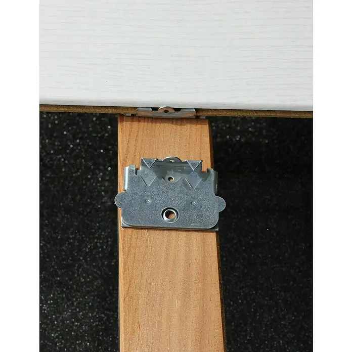 Staba Grapa para friso Inicial - Acero - Primer plano de una fijación metálica unida a un trozo de madera, que sujeta una tabla blanca.