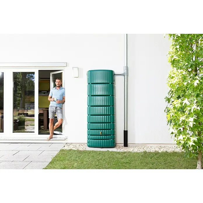 4rain Regenspeicher Slim - 650 l, Grün - Bild eines schlanken, grünen Regenspeichers neben einem Haus mit einem Mann, der in der Tür steht. Eine Regenrinne verläuft daneben.