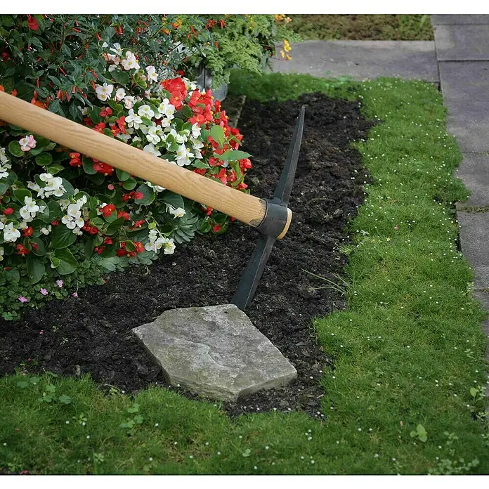 Gardol Kreuzhacke - Material Gerätekopf: Metall, Hackenstiel - Nahaufnahme einer Spitzhacke, die in die Erde gesteckt wird, umgeben von Blumen und Rasen, ideal für Gartenarbeiten.