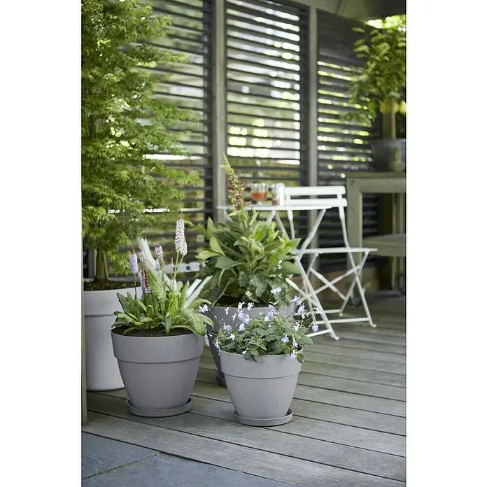 Elho Vibia Campana Maceta para planta Vibia Campana - Dimensiones externas (Ø x Al): 47 x 40,4 cm, Plástico, Gris hormigón - Un primer plano de jardineras grises con flores coloridas en un patio, mostrando una escena de jardín serena y natural.