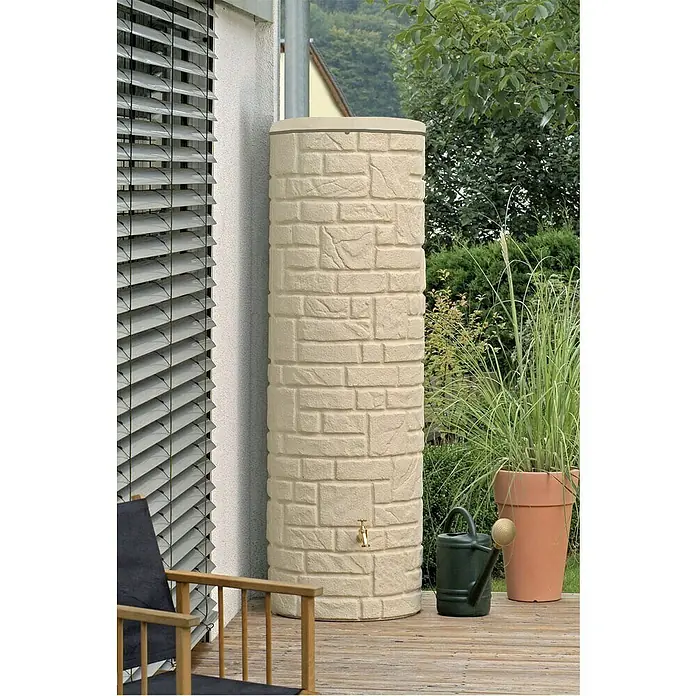 Arves Regenspeicher Arcado - 460 l, Sandsteinfarben - Wassertank in Steinoptik mit Hahn, daneben eine Gießkanne und ein Blumentopf mit hohem Gras auf einer hölzernen Terrasse.