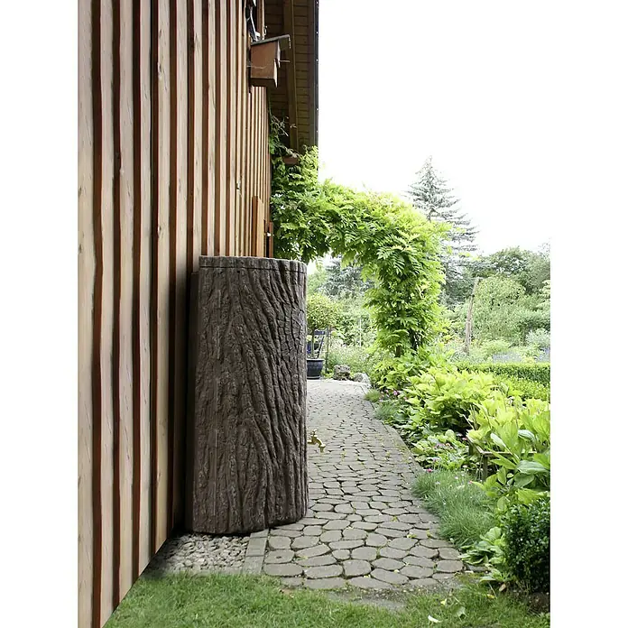 Arves Regenspeicher Evergreen Lite - 300 l, Dunkelbraun - Regentonne in Baumstammoptik neben einem Holzhaus mit grüner Pergola und Steinweg im Garten.