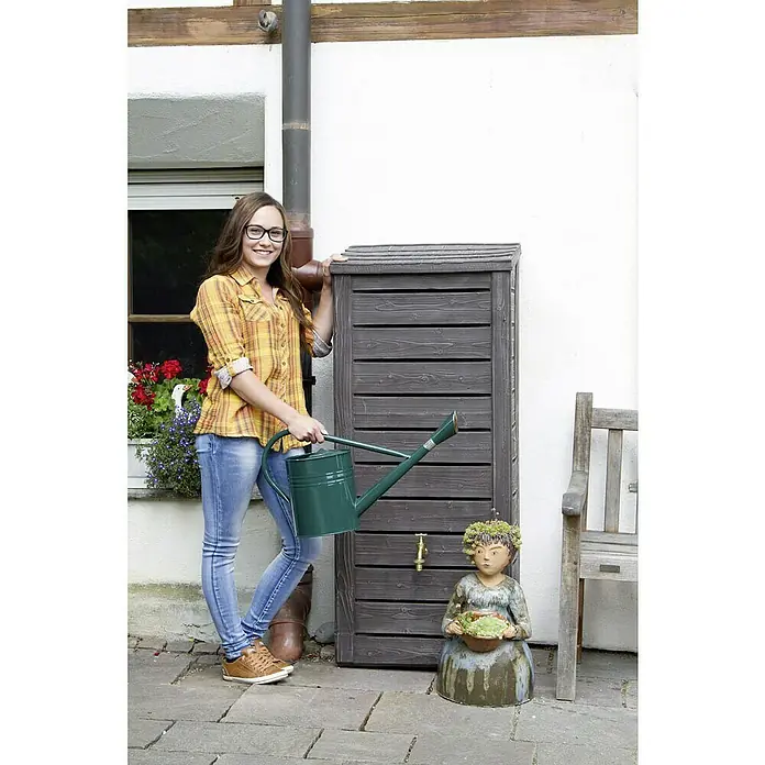 Arves Regenspeicher Norway - 230 l, Dunkelbraun, 1 Stk. - Eine Frau steht neben einem dunklen Holz-Regenwassertank mit einem Wasserhahn. Sie hält eine grüne Gießkanne in der Hand.