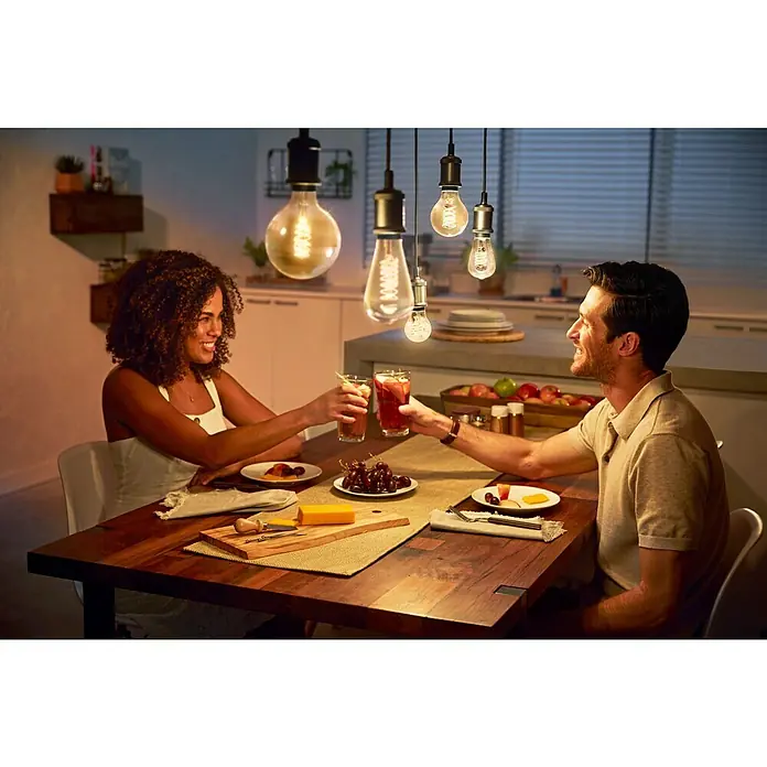 Philips Hue Bombilla LED inteligente Filamento Edison - E27, Intensidad regulable, Blanco cálido, 550 lm, 7 W, Denominación de la lámpara: ST72 - Una pareja brinda con bebidas en una mesa iluminada por bombillas decorativas Philips, creando un ambiente cálido y acogedor.