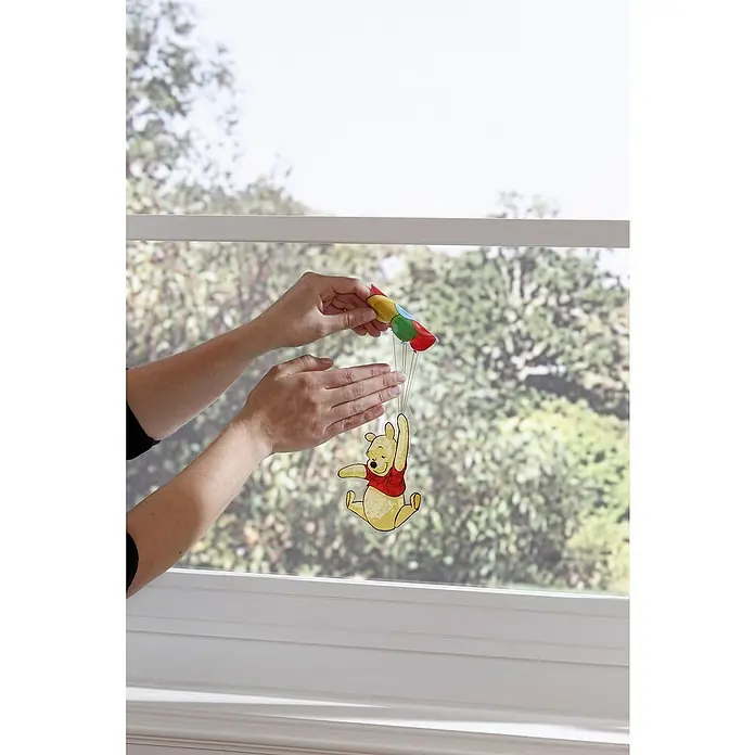 Komar Fenstersticker Winnie Pooh - 31 x 31 cm - Ein Winnie Puuh Fensterbild, das an bunten Ballons hängt, wird von zwei Händen vor ein Fenster gehalten.