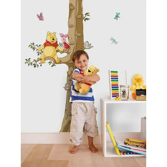 Komar Dekosticker Winnie The Pooh Size - B x H: 100 x 70 cm - Ein Junge umarmt ein Winnie Puh-Plüschtier vor einer Winnie Puh-Wandaufkleber-Größentabelle in einem Kinderzimmer.