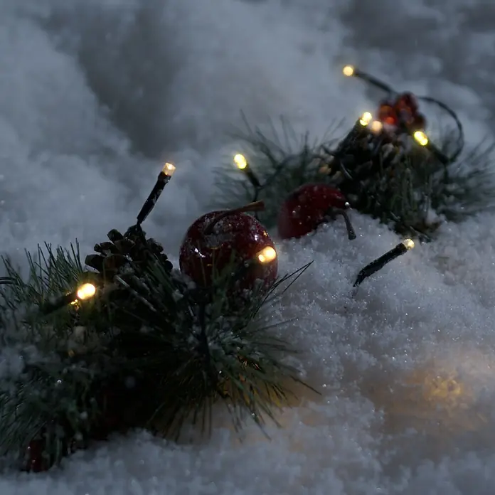 Konstsmide LED-Lichterkette - Außen, Lichtfarbe: Warmweiß, Batteriebetrieben, 0,5 m, Schwarz - Winterliche Szene mit einer Lichterkette, Äpfeln und Tannenzapfen im Schnee, die eine gemütliche und festliche Atmosphäre erzeugt.