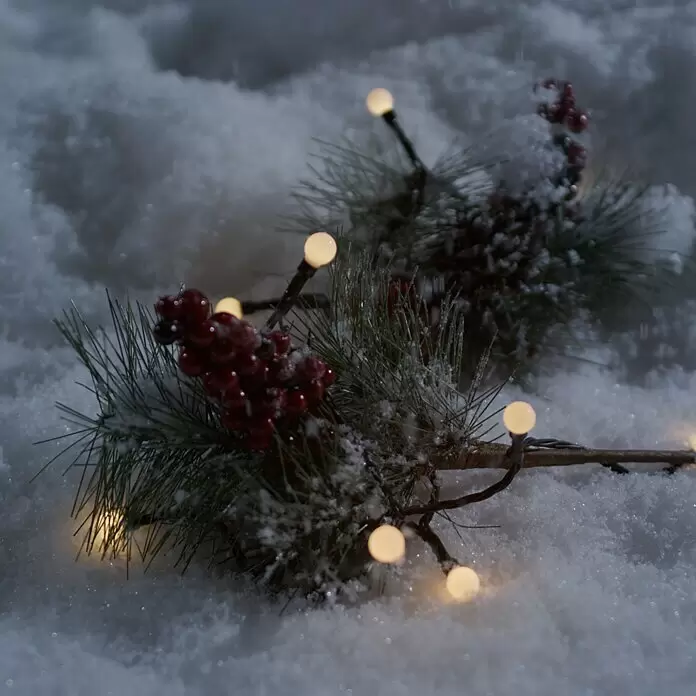 Konstsmide LED-Lichterkette Globes - Außen, Lichtfarbe: Warmweiß, Anzahl Leuchtmittel: 40 Stk., Batteriebetrieben - Eine verschneite Kiefernzweig mit roten Beeren und warmweißen Lichterketten, die eine festliche Winterszene bilden.