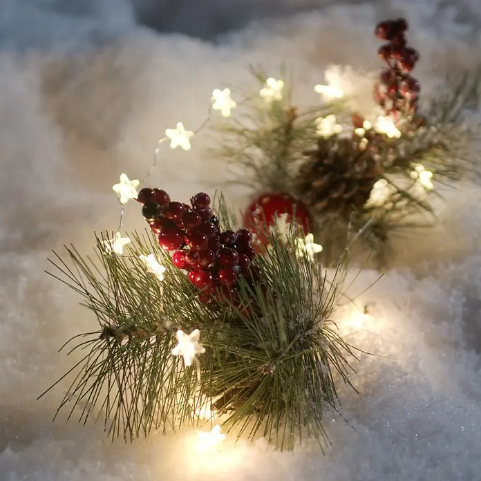 Konstsmide LED-Lichterkette Leuchtsterne - Innen, Lichtfarbe: Warmweiß, Anzahl Leuchtmittel: 20 Stk., Batteriebetrieben - Festliche Weihnachtsdekoration mit Tannenzweigen, roten Beeren und warmen Sternenlichtern auf einer Schneedecke.