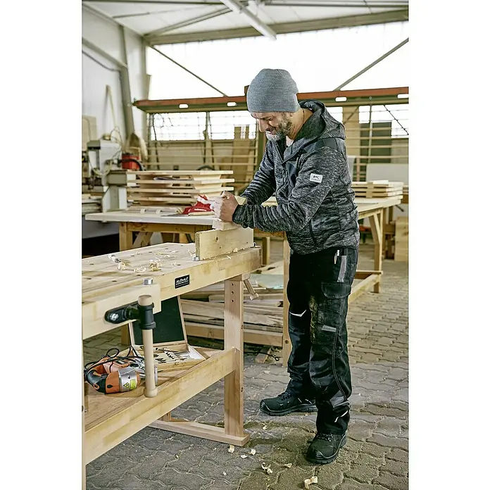 Ramia Hobelbank Advanced 1500 - L x B: 1.500 x 650 mm, Arbeitshöhe: 850 mm, Holz - Ein Handwerker in Arbeitskleidung hobelt ein Holzstück auf einer Werkbank, umgeben von Werkzeugen und Spänen.