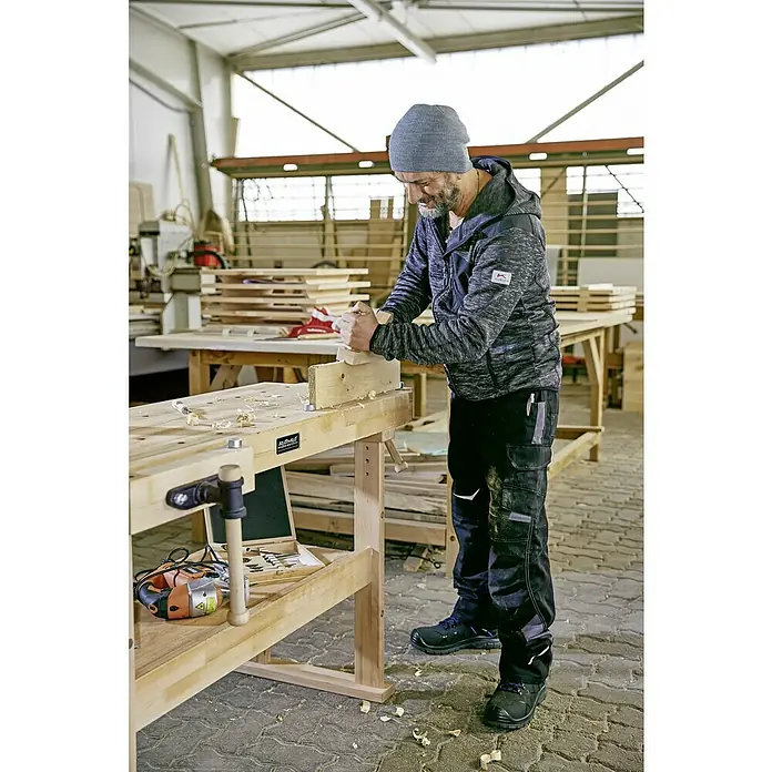 Ramia Hobelbank Advanced 1500 - L x B: 1.500 x 650 mm, Arbeitshöhe: 850 mm, Holz - Ein Handwerker hobelt mit einer Handhobel Holz auf einer Werkbank in einer Werkstatt, bekleidet mit Arbeitskleidung und einer Mütze.