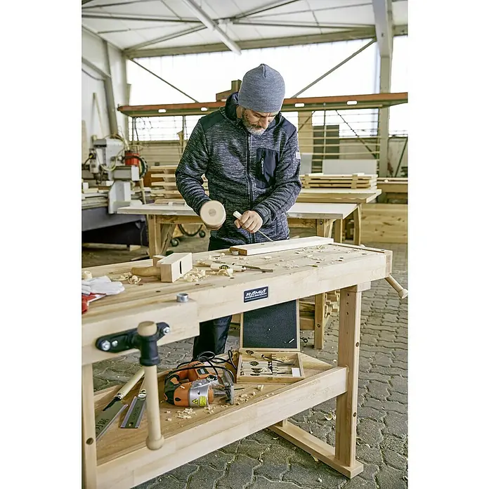 Ramia Hobelbank Advanced 1500 - L x B: 1.500 x 650 mm, Arbeitshöhe: 850 mm, Holz - Ein Handwerker arbeitet mit einem Stecheisen und einem Holzhammer an einer RAMIA Hobelbank. Werkzeuge liegen bereit.