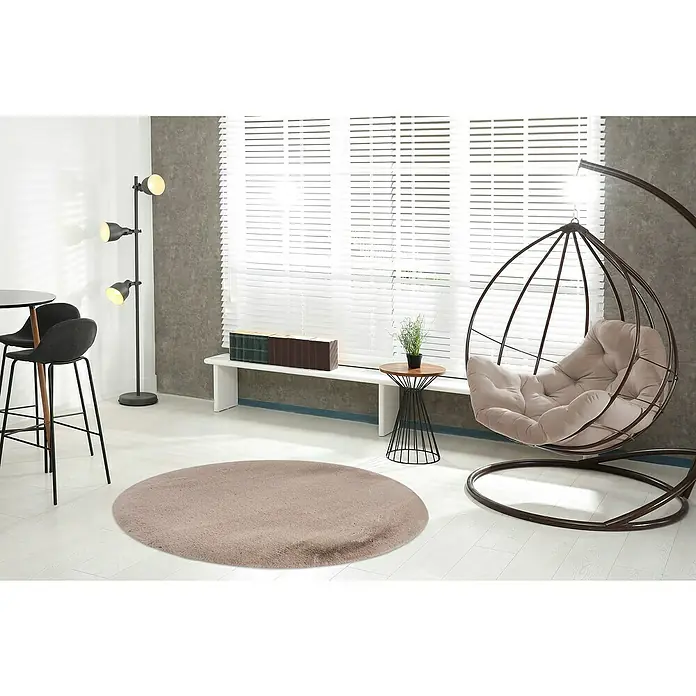 Fellteppich Happy - Taupe, Durchmesser: 160 cm, 100 % Polyester (Flor) - Ein weicher, runder Teppich in sanftem Beigeton liegt in einem stilvollen Wohnzimmer mit Hängekorb und minimalistischer Einrichtung.