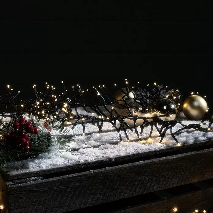 Tween Light LED-Clusterlichterkette 8 Funktionen - Außen, Länge: 12,4 m, 1.000 Stk., Netzbetrieben - Warmweiße LED-Lichterkette mit schwarzem Kabel, drapiert über einer verschneiten Holzkiste mit roten Beeren und goldenen Weihnachtskugeln.