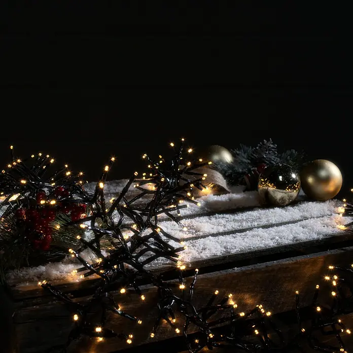 Tween Light Cadena de luces LED - Para exterior, Largo: 12,4 m, 1.000 ud., Funcionamiento en red, Color de luz: Ámbar - Guirnalda de luces decorativa con adornos navideños dorados y ramas de abeto sobre una superficie rústica de madera, ideal para decoraciones festivas.