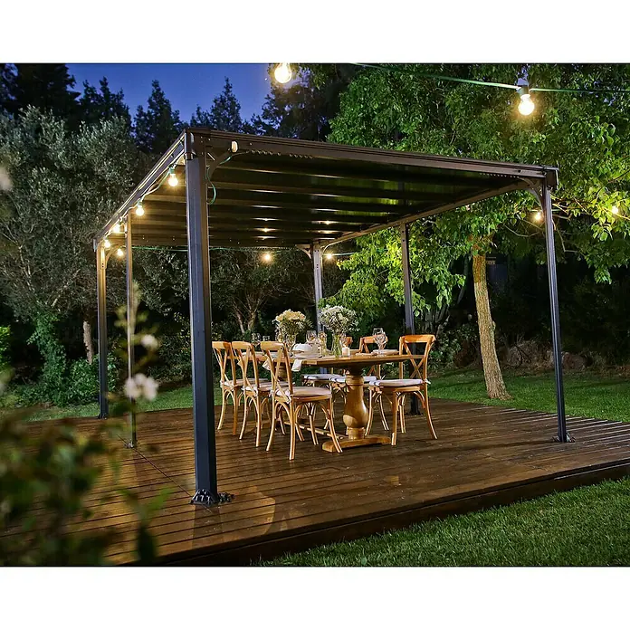 Palram – Canopia Pérgola estándar Milano - An x Pr: 435,9 x 315,5 cm, Color del armazón: Antracita, Aluminio - Pérgola elegante con mesa y sillas en una terraza de madera, iluminada con luces cálidas para un ambiente comedor exterior.