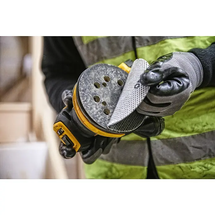 DeWalt XR 18V Akku-Exzenterschleifer DCW210N-XJ - 18 V, Ohne Akku, Durchmesser Schleifteller: 125 mm, Karton - Eine Person befestigt ein Schleifpapier an einem gelben und schwarzen Exzenterschleifer mit Löchern, um Staub zu entfernen. Die Person trägt Handschuhe und eine Warnweste.