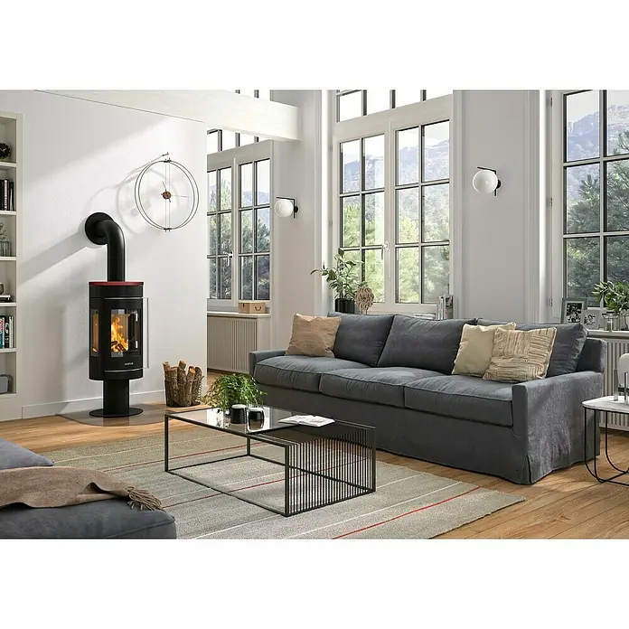 Justus Kaminofen Meva - 5,5 kW, Raumheizvermögen: 88 m³, Farbe Verkleidung: Bordeauxrot, Schwarz - Wohnzimmer mit Justus Kaminofen, grauer Couch und großem Fenster. Der Ofen sorgt für Wärme und Atmosphäre im Raum.