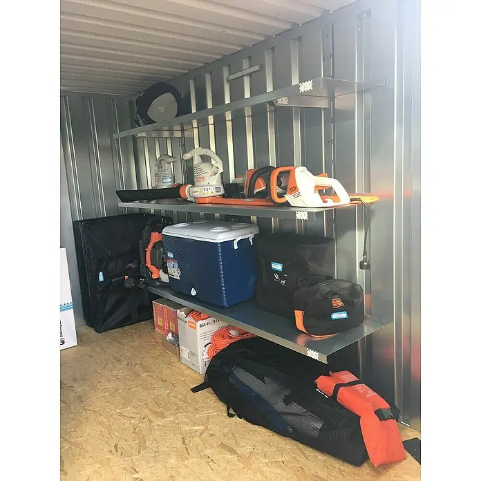 BOS Best of Steel Schnellbaucontainer SC3000+1X2-SE1 x 2,1 x 2,1 m, Stahl, Einzeltür - Lagerraum mit Regalen voller Gartengeräte, darunter Laubbläser, Heckenschere und Zubehör, ordentlich verstaut.
