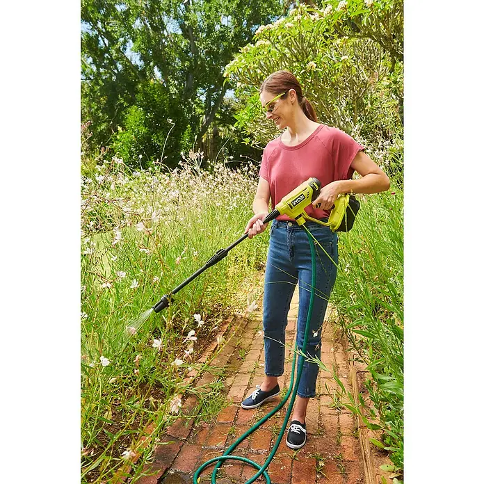 Ryobi ONE+ Akku-Hochdruckreiniger RY18PW22A-125 - 18 V, 1 Akku, 2,5 Ah, Max. Druck: 22 bar, Fördermenge: 182 l/h - Eine Frau benutzt einen Ryobi-Akku-Reiniger im Garten, um den Weg zu säubern, und trägt dabei eine Schutzbrille.
