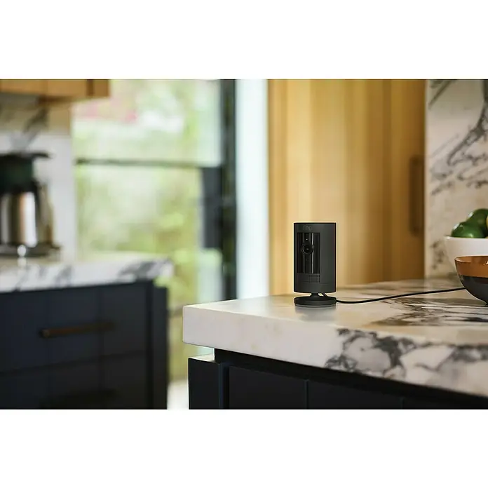 Ring Überwachungskamera außen Stick Up Cam Plug-In - Schwarz, Erfassungswinkel: 110 °, Netzbetrieben - Nahaufnahme einer schwarzen Ring Sicherheitskamera auf einer Küchentheke, die moderne Sicherheitstechnologie zeigt.