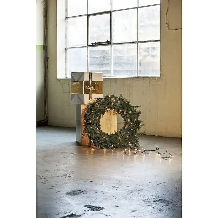Konstsmide LED-Lichterkette - Innen, Lichtfarbe: Bernstein, Anzahl Leuchtmittel: 50 Stk., Netzbetrieben - Festliche Dekoration mit einem goldfarbenen Geschenk, einem Kranz mit Lichtern und einer Lichterkette auf einem zementfarbenen Boden vor einem Fenster.