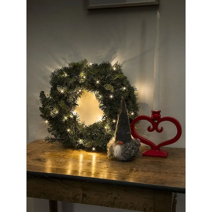 Konstsmide LED-Lichterkette - Innen, Lichtfarbe: Bernstein, Anzahl Leuchtmittel: 50 Stk., Netzbetrieben - Festliche Dekoration mit einem beleuchteten Weihnachtskranz, einem Zwerg und einem Kerzenständer in Herzform auf einem Holztisch.