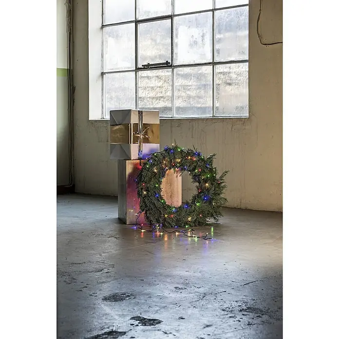 Konstsmide LED-Lichterkette - Innen, Lichtfarbe: Bernstein, Anzahl Leuchtmittel: 50 Stk., Netzbetrieben - Festliche Szene mit Geschenkstapel und einem beleuchteten Weihnachtskranz vor einem großen Fenster, ideal für saisonale Dekorationen.