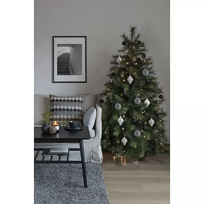 Konstsmide Baummantel - Innen, 2,4 m, 5 Stränge à 40 gefrostete bernsteinfarbene LEDs - Grüner künstlicher Weihnachtsbaum, geschmückt mit weißen Lichterketten und schwarz-weißen geometrischen Papierornamenten, neben einem Sofa.