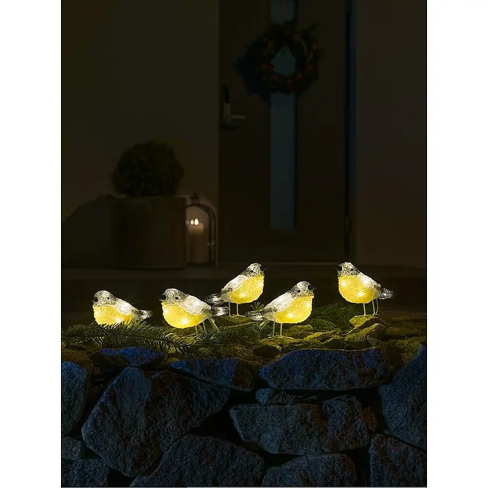 Konstsmide LED-Weihnachtsdekoleuchte Acryl Vögel - 4 m x 16 cm x 10 cm, Klar, Netzbetrieben, LED - Bild von einer Gruppe von leuchtenden Deko-Vögeln, die auf einer Steinmauer sitzen, mit festlicher Dekoration im Hintergrund, ideal für stimmungsvolle Beleuchtung.