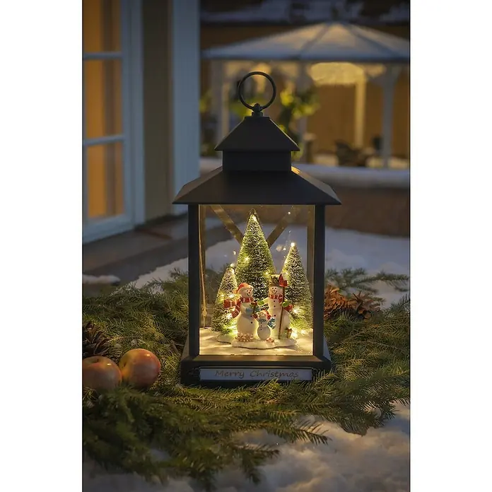 Konstsmide LED-Weihnachtsdekoleuchte - 24,5 x 24,5 x 42 cm, Schwarz, Netzbetrieben, LED - Festliche Weihnachtslaterne mit Schneemannfamilie und funkelnden Lichtern, umgeben von Tannengrün und Äpfeln, die eine warme, winterliche Atmosphäre schafft.