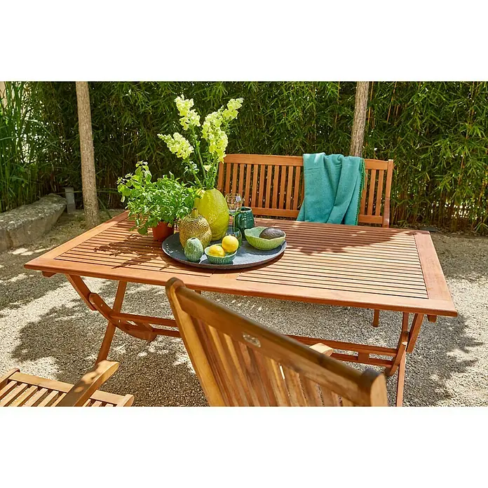 Sunfun Diana Gartentisch - L x B x H: 150 x 86 x 75 cm, Akazie, Rechteckig, Naturbraun - Ein hölzerner Gartentisch mit Dekorationen und Stuhl im Vordergrund vor einem üppigen grünen Hintergrund.