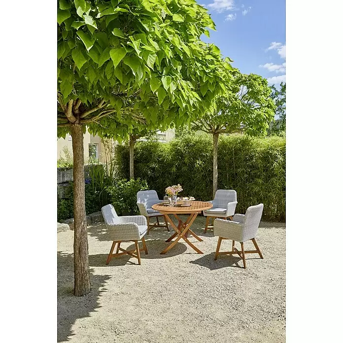 Sunfun Diana Gartentisch rund - Ø x H: 107 x 75 cm, Akazie, Naturbraun, Klappbar - Runder Gartentisch aus Holz mit vier grauen Korbstühlen und hellen Kissen auf einem Kiesweg unter Bäumen mit Snacks.