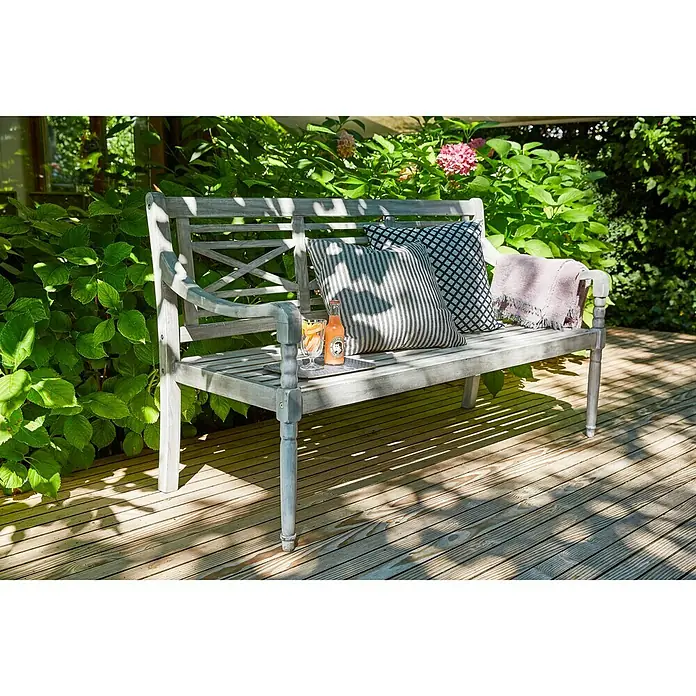Sunfun Gartenbank Alea - B x T x H: 150 x 62 x 87 cm, Akazie, White Washed - Eine graue Gartenbank mit Kissen und Getränken auf einer Holzterrasse, umgeben von üppigem Grün. Perfekt zum Entspannen im Freien.