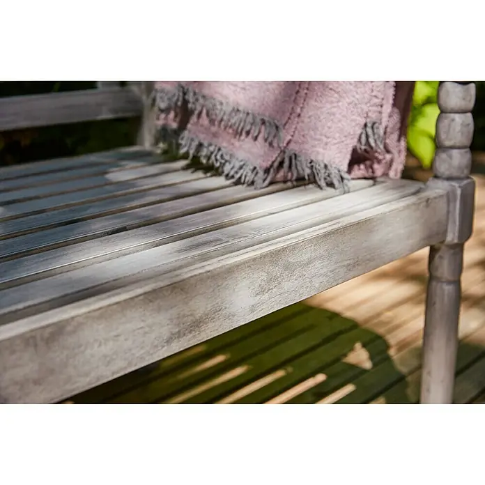 Sunfun Gartenbank Alea - B x T x H: 150 x 62 x 87 cm, Akazie, White Washed - Nahaufnahme einer grauen Gartenbank mit einer rosafarbenen Decke. Die Bank steht auf einer Holzterrasse und ist von Schatten umgeben.