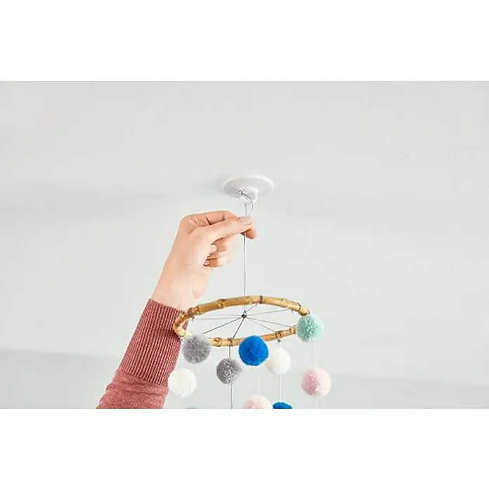 Tesa Colgador adhesivo de techo - Blanco - Una persona sujeta un móvil de pompones colorido a un gancho del techo, creando un elemento lúdico y decorativo.