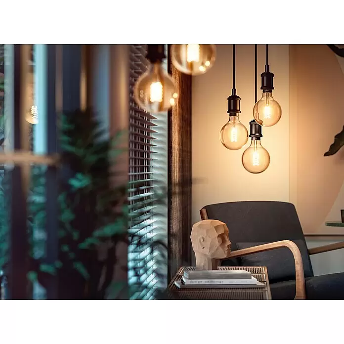 Philips Hue Bombilla LED inteligente Filamento blanco - E27, Intensidad regulable, Blanco cálido, 550 lm, 7 W, Denominación de la lámpara: G93 - Tres bombillas colgantes de cristal ámbar con filamentos en espiral brillantes en un ambiente interior acogedor.