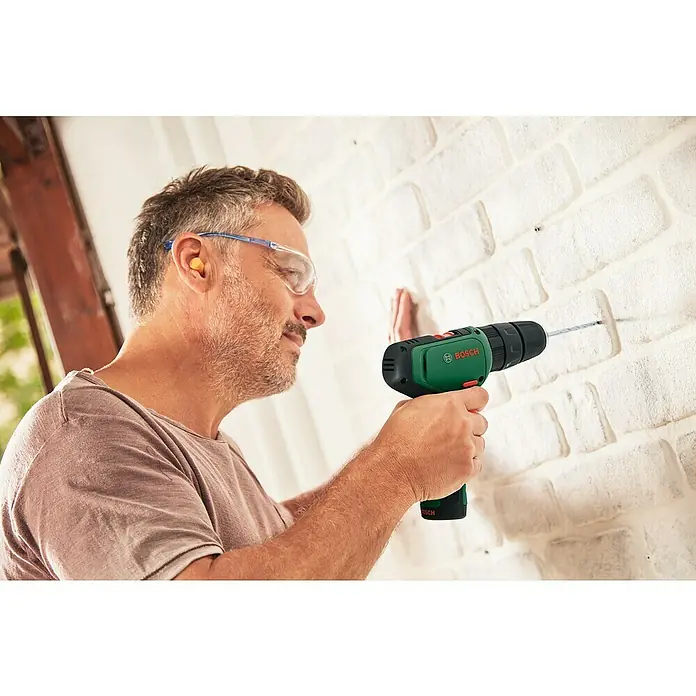 Bosch Power for All 12V Akku-Schlagbohrschrauber EasyImpact 1200 - 12 V, 1 Akku, 1,5 Ah - Ein Mann bohrt mit einem grünen Bosch Akku-Bohrschrauber ein Loch in eine weiße Ziegelwand, während er Schutzbrille und Ohrenstöpsel trägt.
