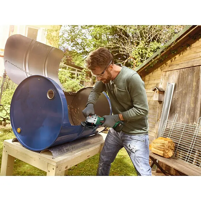Bosch Power for All 18V Amoladora angular de batería AdvancedGrind 18 - 18 V, Sin batería, Diámetro disco: 125 mm - Un hombre utiliza una amoladora angular en un barril azul al aire libre, el barril se está utilizando para un proyecto de barbacoa.