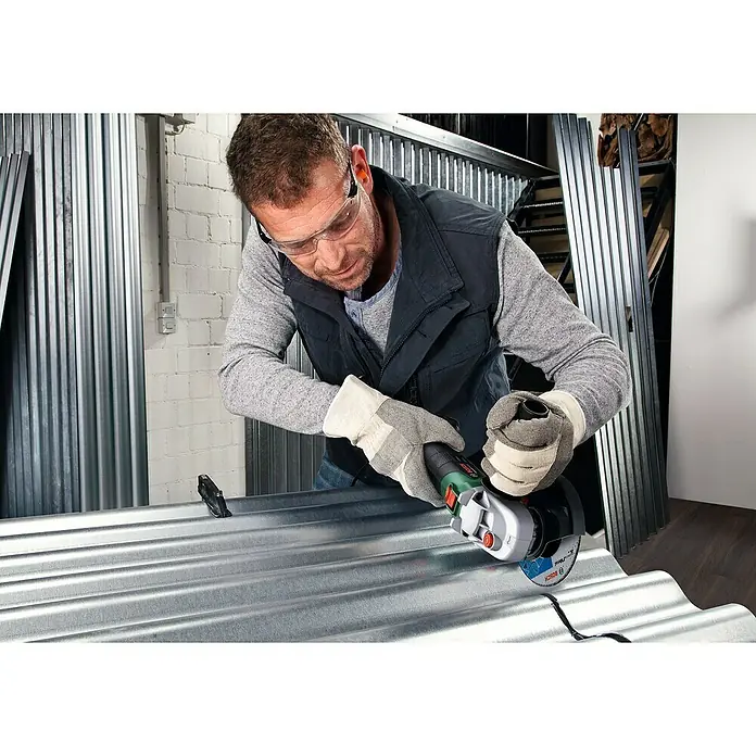 Bosch Power for All 18V Amoladora angular de batería AdvancedGrind 18 - 18 V, Sin batería, Diámetro disco: 125 mm - Hombre con gafas de seguridad que utiliza una amoladora angular Bosch para cortar una pieza de metal corrugado mientras usa guantes.