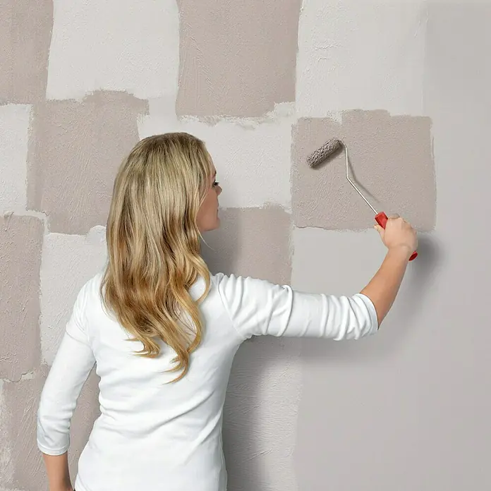 SCHÖNER WOHNEN-Farbe Trendstruktur Dekoputz-Set Loft-Optik - 5 -tlg. - Eine Frau streicht eine Wand mit einem Farbroller in einem warmen Grauton, um ein modernes, gestreiftes Design zu schaffen.