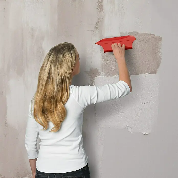 SCHÖNER WOHNEN-Farbe Trendstruktur Dekoputz-Set Loft-Optik - 5 -tlg. - Frau glättet eine Wand mit einem roten Werkzeug, um eine ebene Oberfläche zu schaffen. Ideal für Renovierungsarbeiten.