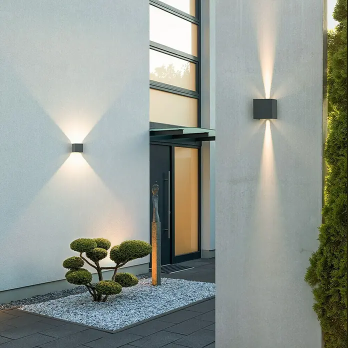 Starlux LED-Außenwandleuchte Umea - 8 W, 10 x 10 x 10 cm, Anthrazit, IP54 - Moderne Außenwandleuchten mit Lichtkegeln beleuchten Hauswand neben einer Tür und einem dekorativen Baum.