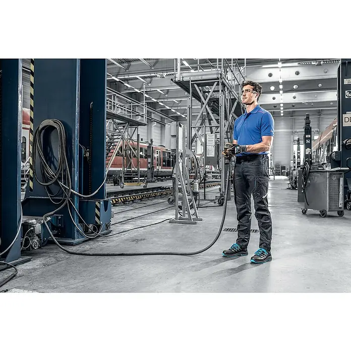 Uvex 2 Trend Sicherheitshalbschuh S3 - 46, Schutzklasse nach EN ISO 20345: S3, Schwarz/Blau - Der Mann trägt blaue Arbeitskleidung und Sicherheitsschuhe in einer industriellen Umgebung mit Werkzeugen und Zügen im Hintergrund.
